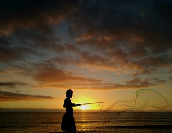 Silhouette man making large bubbles at beach against orange sky during sunset