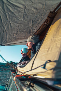 Male hiker in warm clothes lying on blanket in camping tent and drinking cup of coffee in morning and admiring nature at dawn