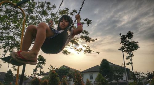 Low angle portrait of girl swinging in park against sky during sunset