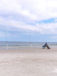 Full length of man with fishing rods sitting at beach against sky