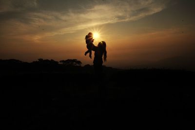 Silhouette woman picking up daughter against sky during sunset