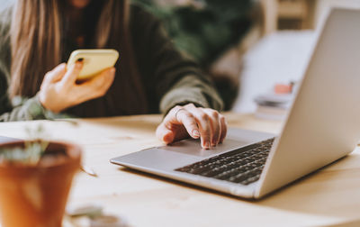 Freelance woman with mobile phone typing at laptop and working from home office with plants. 