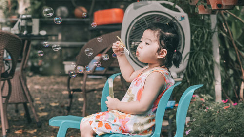 Full length of cute girl blowing bubbles while sitting on chair at yard