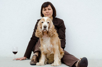 Portrait of woman with cocker spaniel by wineglass sitting against wall