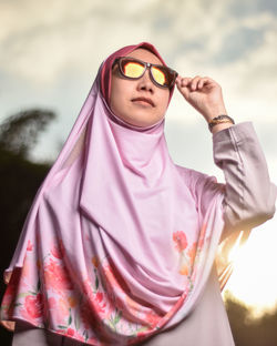 Low angle view of young woman in hijab wearing sunglasses while standing against sky during sunset
