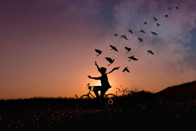 Silhouette child on bicycle reaching birds against sky during sunset