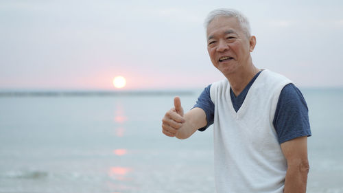 Portrait of smiling man showing thumbs up while standing at beach during sunset