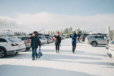 Female and male friends holding skis while walking on snow at parking lot against sky