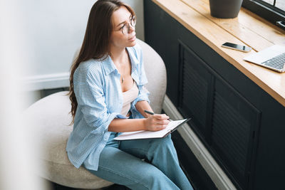 Young woman in bue shirt with notes in hands sitting on chair in room