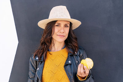 Portrait of woman wearing hat holding fruit against wall