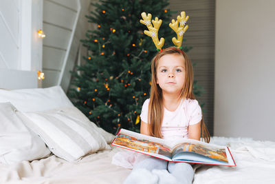 Cute little girl in pink dress reading book sitting on bed in room with christmas tree