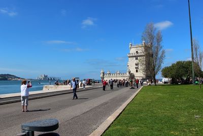 People on footpath leading towards historic building against blue sky