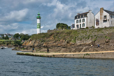 Lighthouse amidst buildings by sea against sky