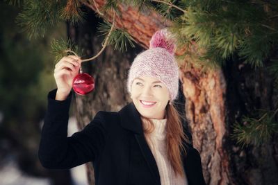 Smiling woman holding bauble by tree during winter