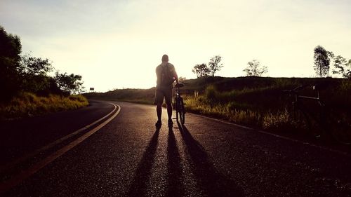 Rear view of man walking with bicycle on country road against sky