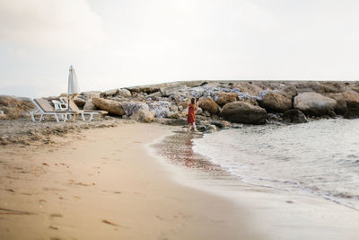 Cute long-haired european baby girl in a dress on the sea coast. girl and stones at sunset on ocean.