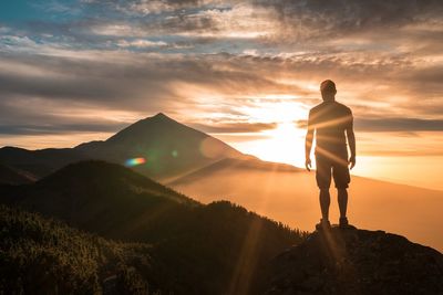 Rear view of mid adult man standing on rock against cloudy sky during sunset