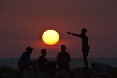 Silhouette people sitting on rock by sea against sky during sunset