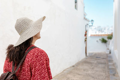 Side view of cheerful female traveler in straw hat on walkway between old buildings in white village in rhodes