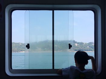 Rear view of boy looking through window while traveling in boat on sea