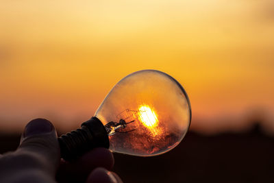 Close-up of cropped hand holding light bulb against sky during sunset