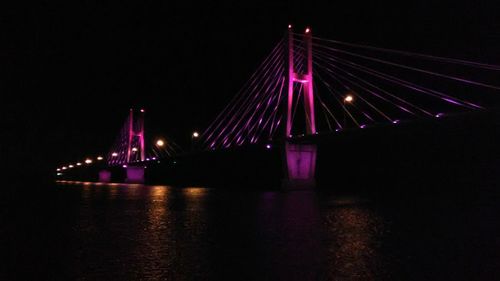 Illuminated suspension bridge over river against sky at night