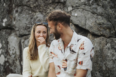 Happy woman talking to male friend while sitting near rocks