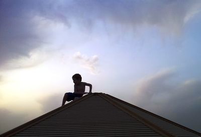 Low angle view of boy on roof against cloudy sky