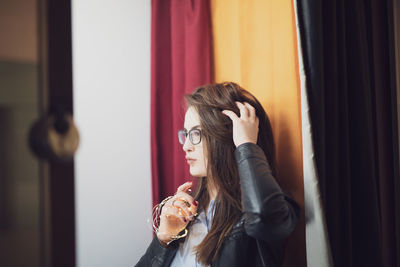 Young woman with hand in hair looking away while standing against curtains at home