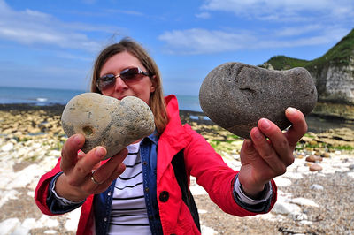 Portrait of smiling young man holding sunglasses against sky