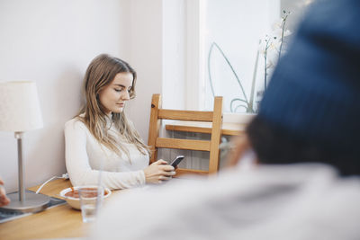 Woman using mobile phone sitting at table against wall in dorm