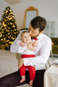 A stylish dad in a white shirt with a red bow tie holds his son in his arms against the background