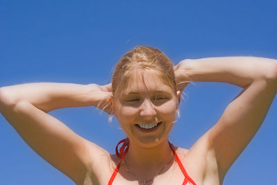 Smiling young woman with hands behind head against clear blue sky