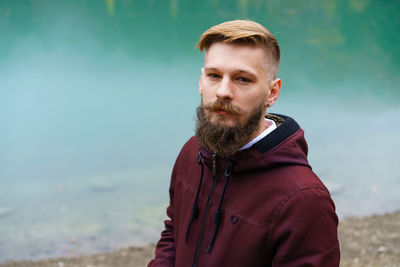 Young bearded man stood alone by lake and looked aside thoughtfully smoking