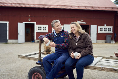 Couple holding mugs while sitting on old-fashioned trailer against barn