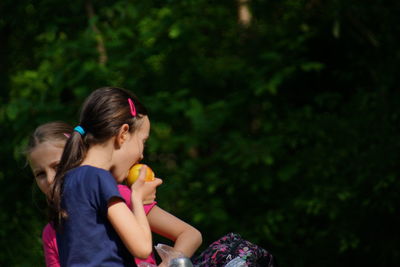 Girl eating apple by sister against plant