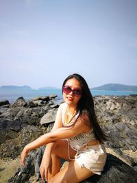 Woman wearing sunglasses while sitting on rock at beach against sky