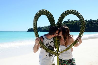 Couple kissing with heart shape patterned palm leaves at beach against sky