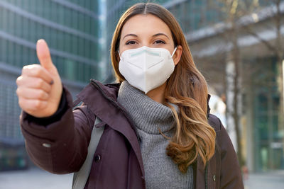 Woman wearing protective mask kn95 ffp2 showing thumbs up in modern city street and looking camera