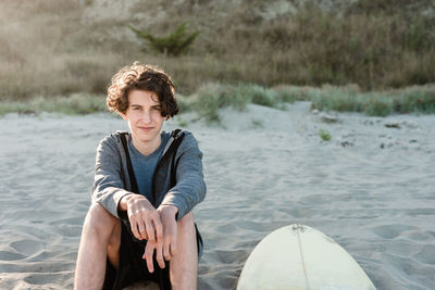 Long haired teenager sitting next to surfboard on a beach in new zealand