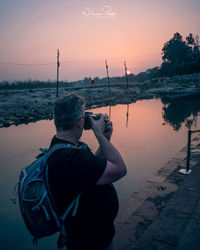 Rear view of man photographing by river against sky