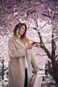 Young woman standing under cherry blossom and using phone