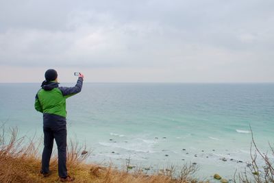 Man in green waterproof jacket takes photo of coastliny by smart phone. cold windy day above sea.