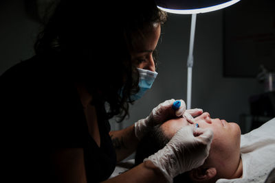 Side view of cosmetician in latex gloves applying purple chemical solution on eyelid of female client during eyelashes lifting procedure in salon