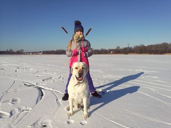 Portrait of girl holding cattail with dog on snow covered field against clear blue sky