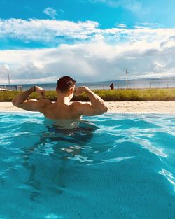 Rear view of shirtless man flexing muscles in swimming pool by sea against sky