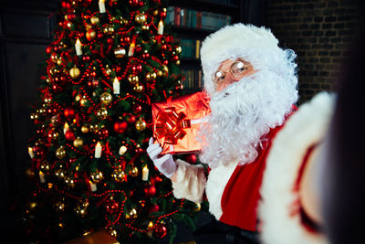 Portrait of santa clause holding gift by christmas tree at home