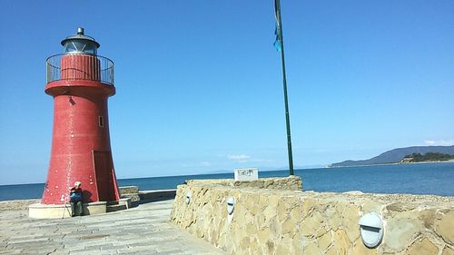 Lighthouse by sea against clear blue sky