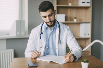 Friendly young male doctor of oriental appearance sits at a desk in his office and takes notes 