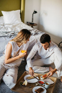 From above of happy young couple sitting together near bed and communicating while enjoying breakfast with pastry toasts and beverages during honeymoon